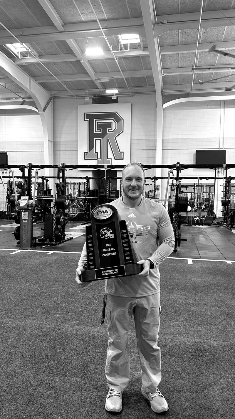 Scott Leech holding the 2024 CAA Football Champions trophy in the URI weight room